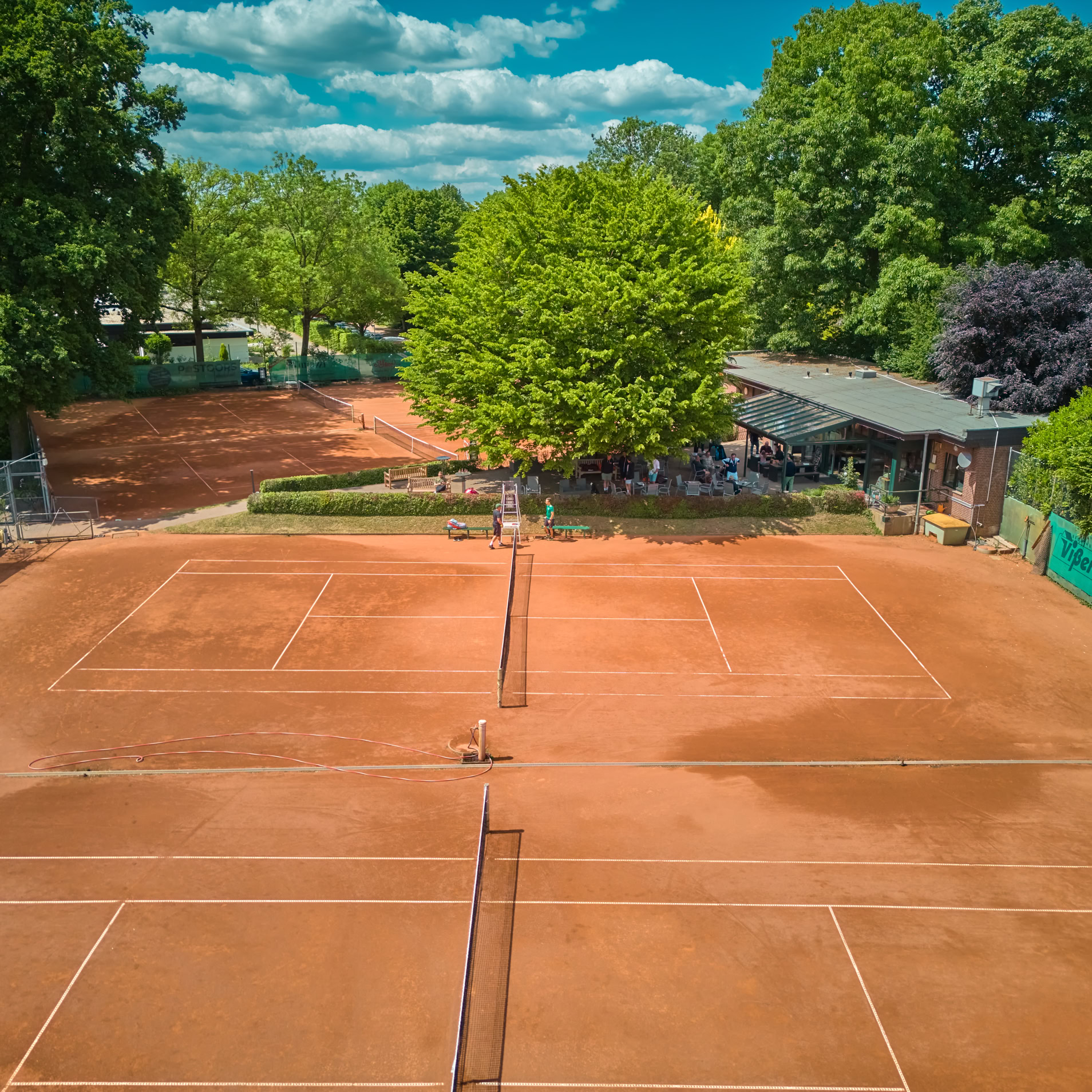 Tennisplatz mit rotem Sandbelag, umgeben von B&auml;umen und einem Geb&auml;ude im Hintergrund.