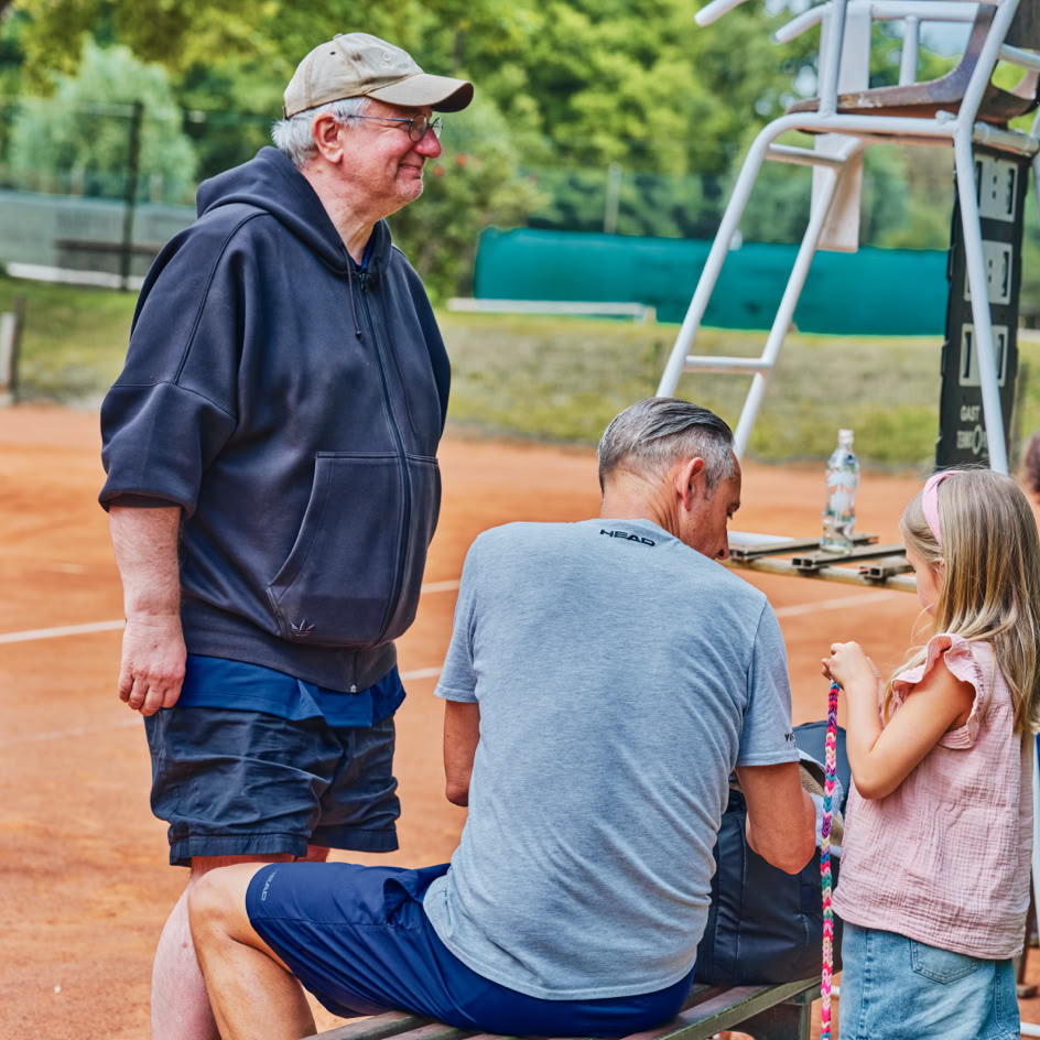 Eine Gruppe von Menschen auf einem Tennisplatz; ein Mann steht, w&auml;hrend ein anderer sitzt und mit einem M&auml;dchen spricht.