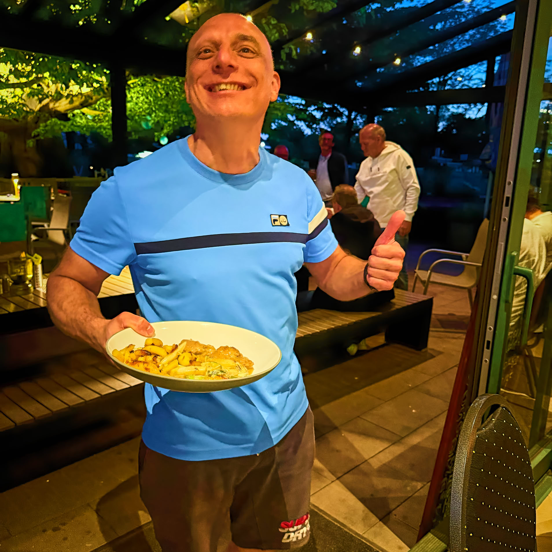 Mann in blauem T-Shirt h&auml;lt einen Teller mit Essen und zeigt Daumen hoch, im Hintergrund ein beleuchteter Au&szlig;enbereich mit Pflanzen und einer weiteren Person.