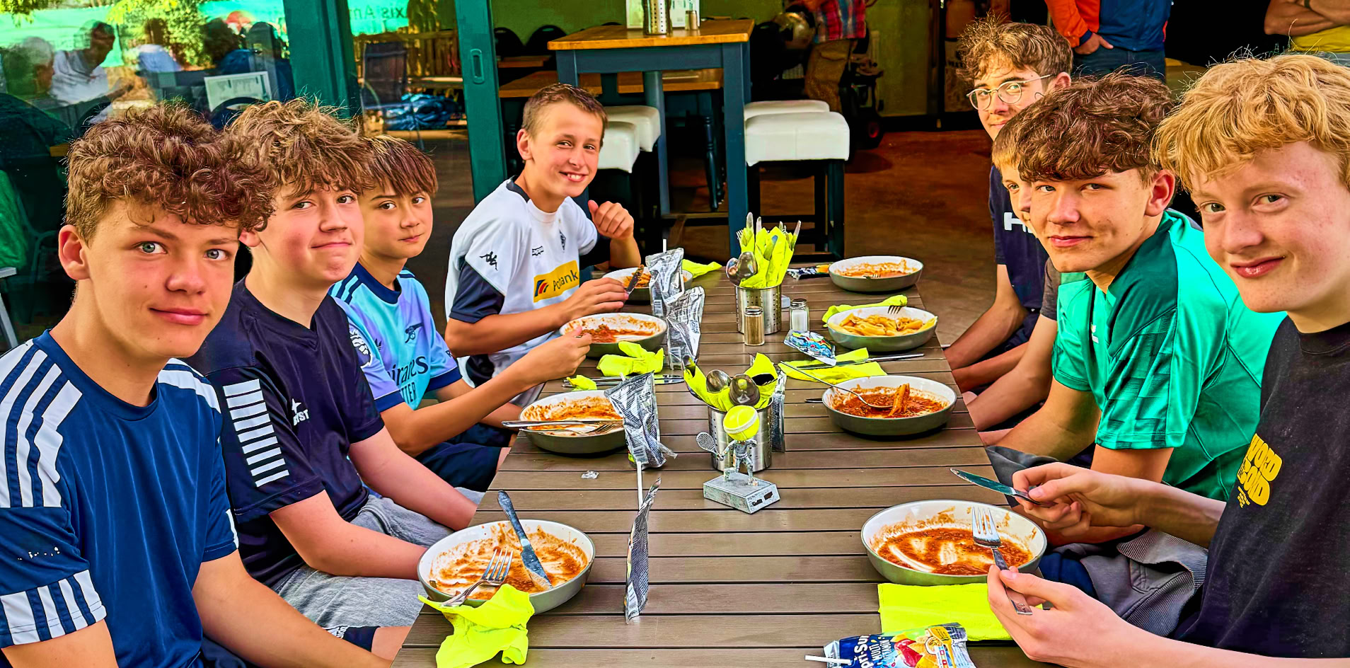 Eine Gruppe von sieben Jugendlichen sitzt an einem Tisch im Freien und isst.