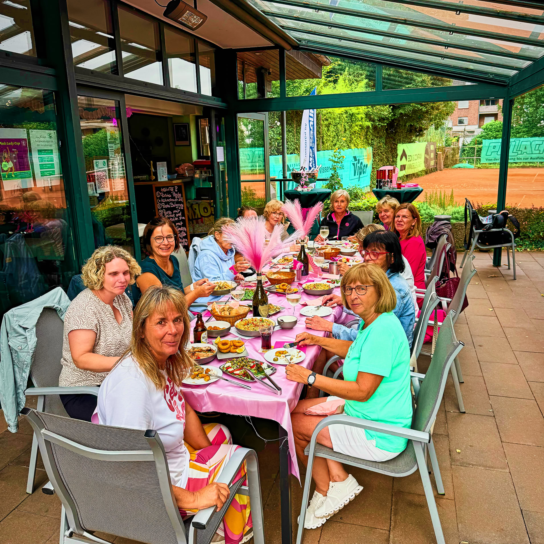 Eine Gruppe von Menschen sitzt an einem langen Tisch im Freien unter einem Glasdach, mit Essen und Getr&auml;nken auf dem Tisch. Einige tragen bunte H&uuml;te.