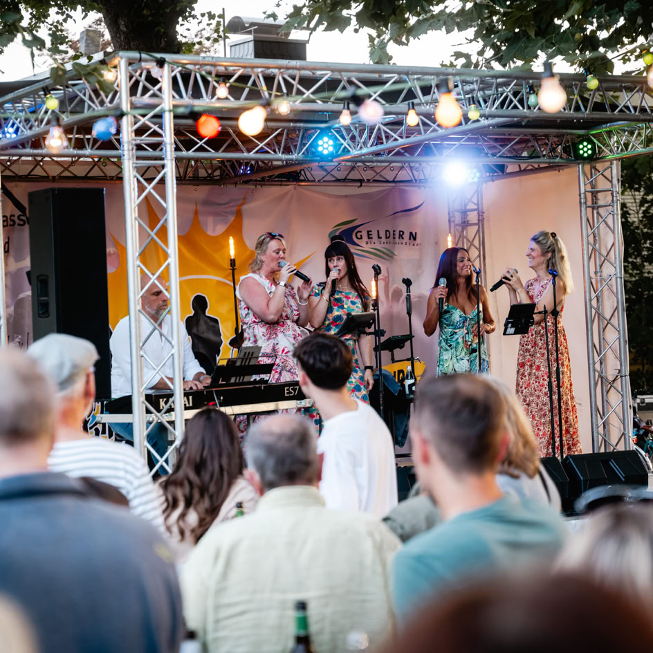 Eine Gruppe von Menschen steht auf einer B&uuml;hne und singt, w&auml;hrend ein Publikum im Vordergrund zuschaut. Die B&uuml;hne ist mit bunten Lichtern dekoriert.