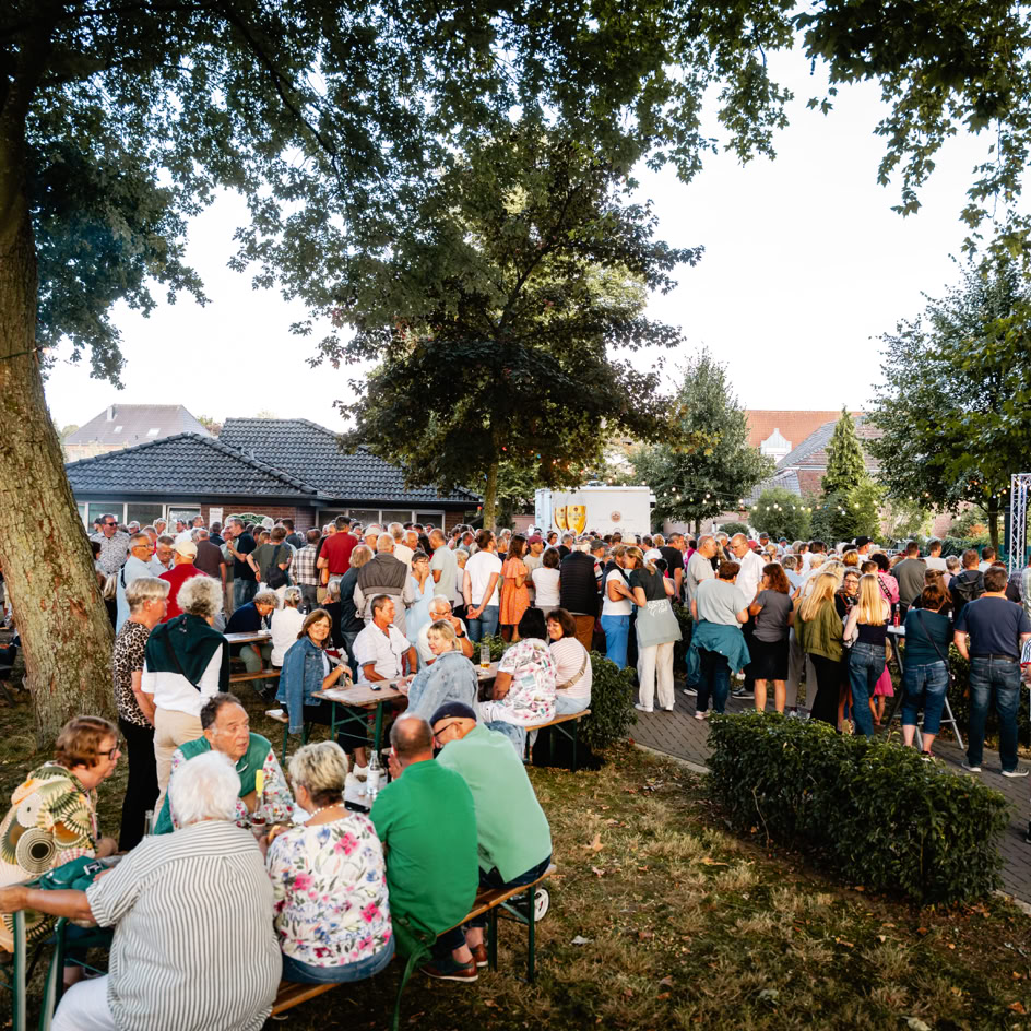 Eine gro&szlig;e Gruppe von Menschen versammelt sich im Freien unter B&auml;umen, einige sitzen an Tischen, w&auml;hrend andere stehen. Im Hintergrund sind Geb&auml;ude und St&auml;nde zu sehen.