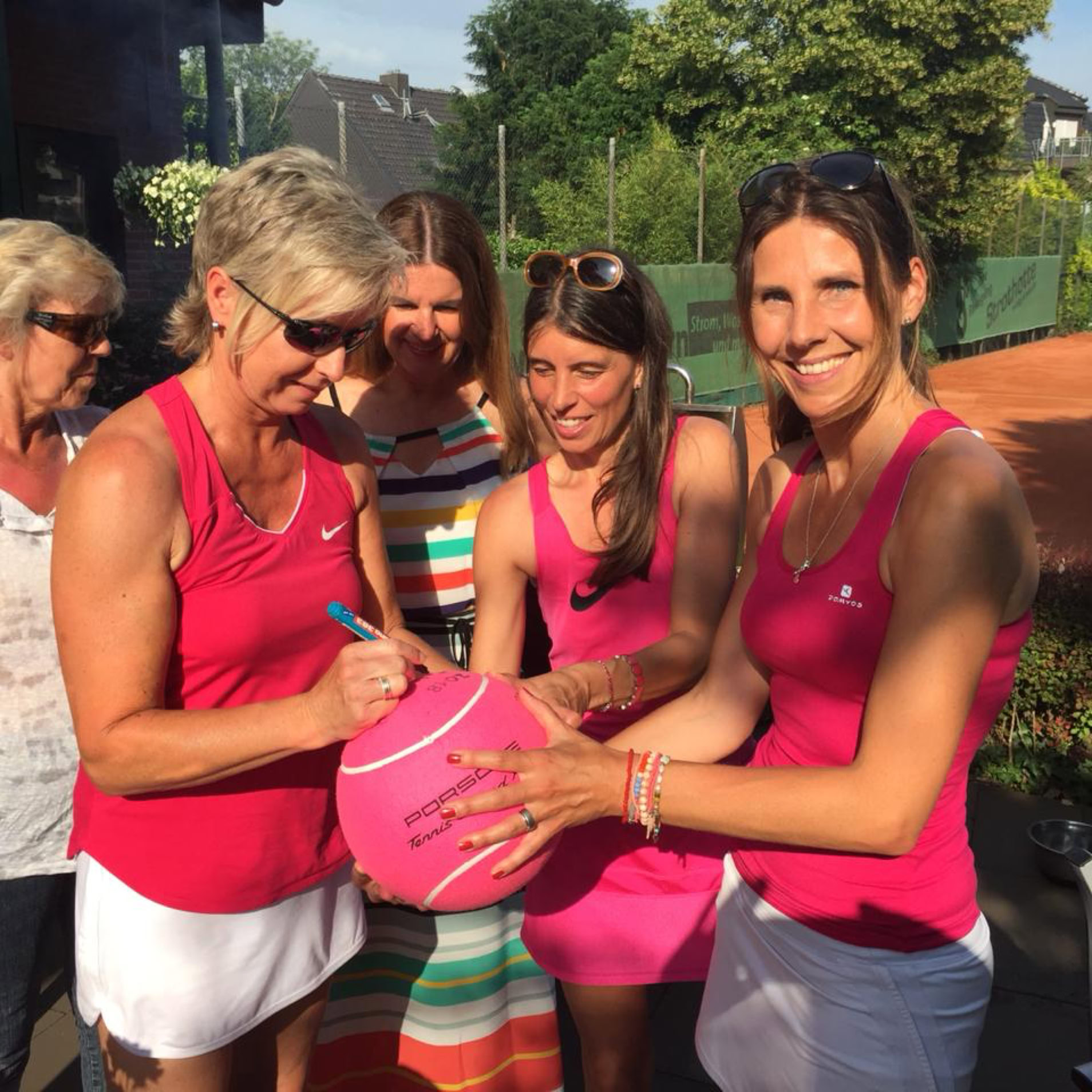 F&uuml;nf Frauen in Sportkleidung, drei tragen pinke Oberteile, stehen zusammen; eine Frau signiert einen gro&szlig;en pinken Tennisball. Im Hintergrund ist ein Tennisplatz zu sehen.