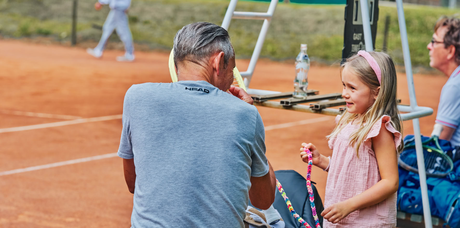 Ein Mann sitzt auf einer Bank auf einem Tennisplatz, neben ihm steht ein M&auml;dchen. Im Hintergrund ist ein Schiedsrichterstuhl und ein weiterer Mann zu sehen.