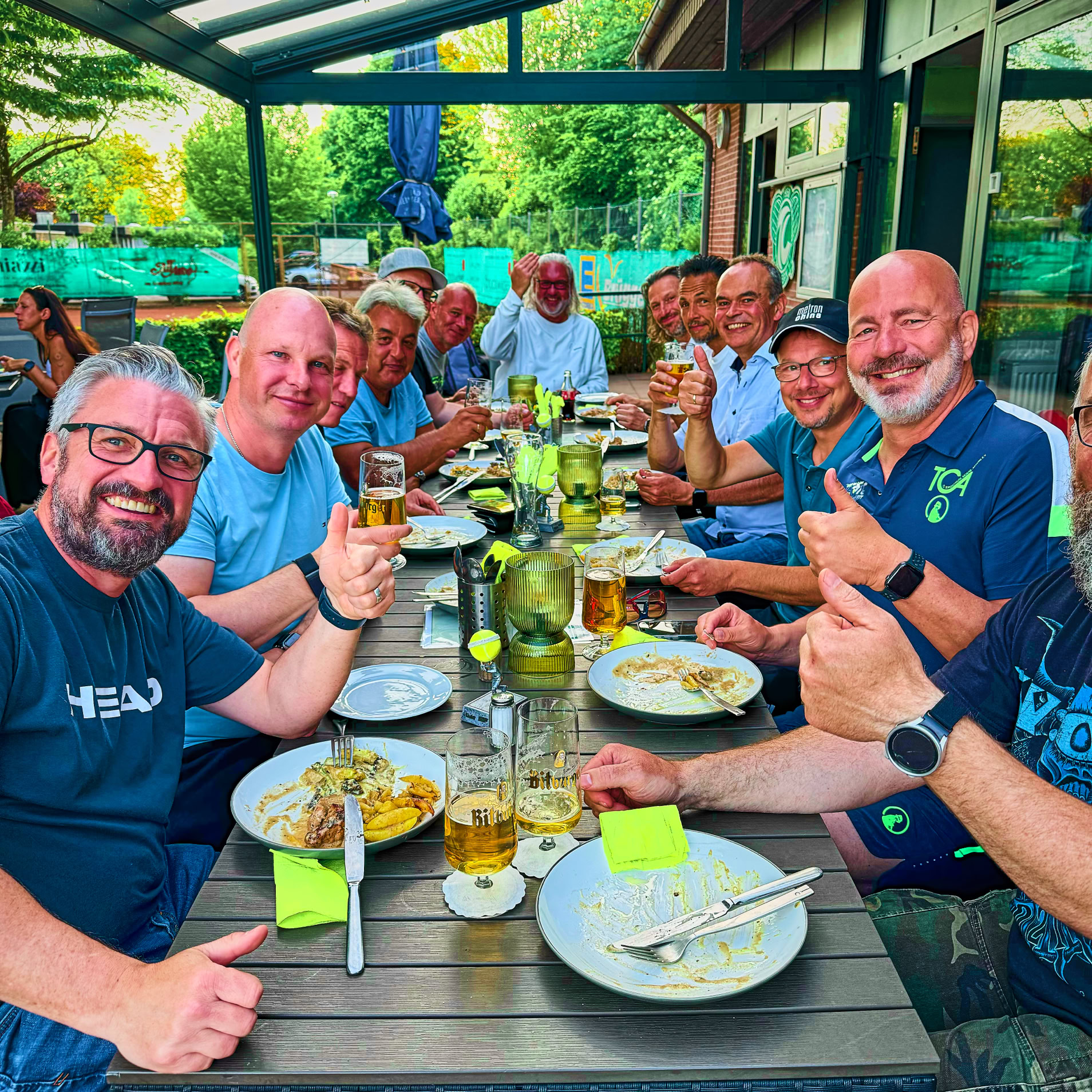 Gruppe von Menschen sitzt an einem Tisch im Freien unter einem Glasdach, isst und trinkt, mit Tellern und Gl&auml;sern auf dem Tisch.