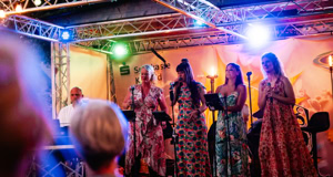 Eine Gruppe von f&uuml;nf Personen steht auf einer B&uuml;hne unter bunten Lichtern, vier davon singen in Mikrofone, w&auml;hrend eine Person ein Keyboard spielt.