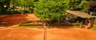 Zwei Sandtennispl&auml;tze umgeben von gr&uuml;nen B&auml;umen und einem Geb&auml;ude im Hintergrund.