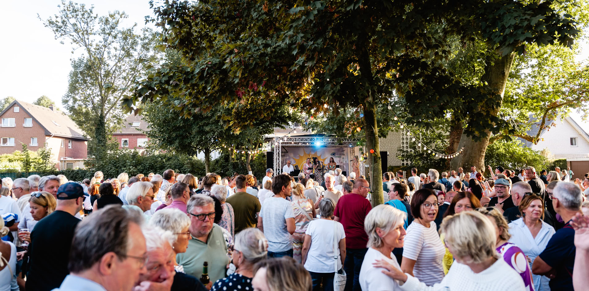 Eine gro&szlig;e Menschenmenge versammelt sich im Freien unter B&auml;umen, einige Personen unterhalten sich, w&auml;hrend andere in Richtung einer B&uuml;hne blicken. Im Hintergrund sind H&auml;user und B&auml;ume zu sehen.