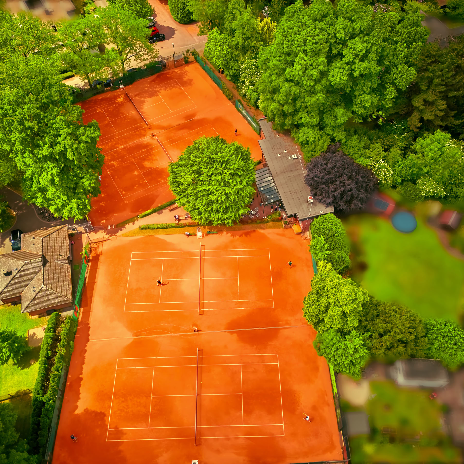 Luftaufnahme von zwei roten Tennispl&auml;tzen, umgeben von gr&uuml;nen B&auml;umen und Geb&auml;uden.