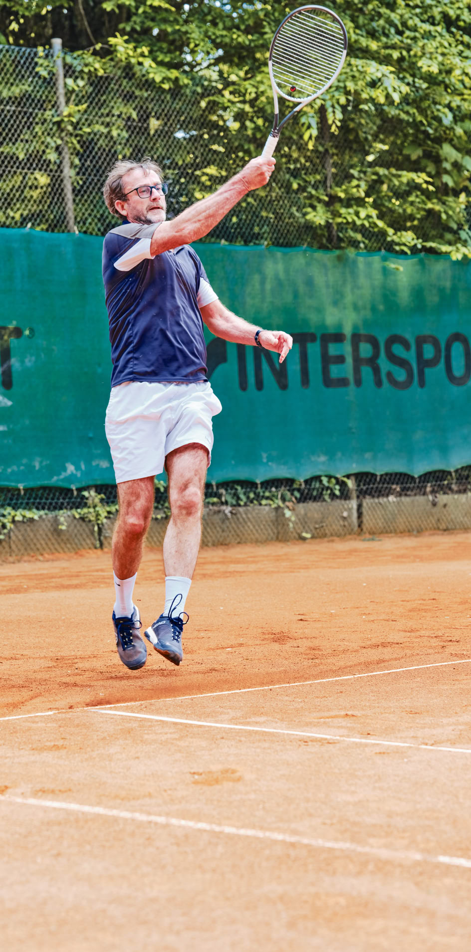Ein Tennisspieler in blauer Kleidung und wei&szlig;en Shorts springt, um einen Ball auf einem Sandplatz zu schlagen, mit einem gr&uuml;nen Zaun im Hintergrund.