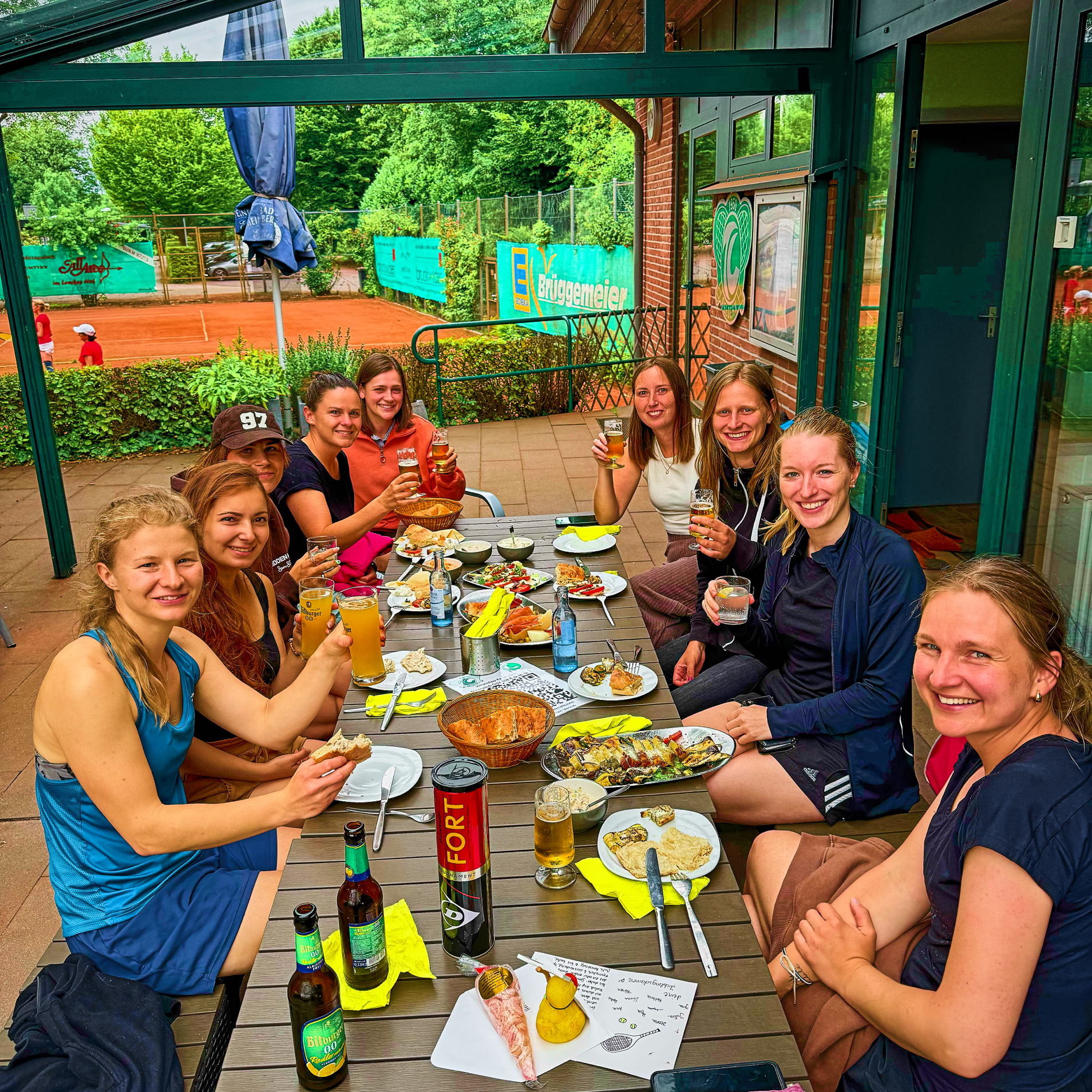 Eine Gruppe von Menschen sitzt an einem Tisch im Freien und isst, mit Getr&auml;nken und verschiedenen Speisen auf dem Tisch. Im Hintergrund sind B&auml;ume und ein Tennisplatz zu sehen.