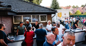 Menschen stehen in einer Schlange vor einem Geb&auml;ude mit einem dunklen Dach, w&auml;hrend andere im Hintergrund auf einem Platz versammelt sind.