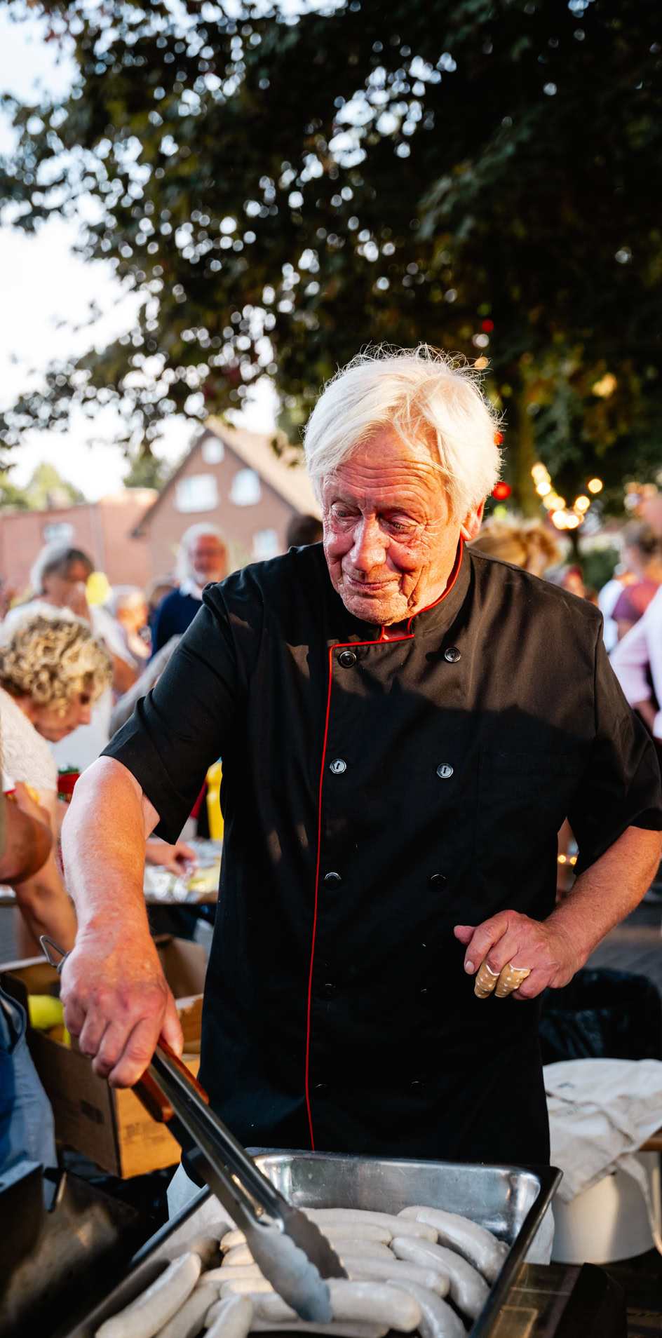 Mann in schwarzer Kochkleidung grillt W&uuml;rste im Freien, umgeben von Menschen bei einer Veranstaltung.