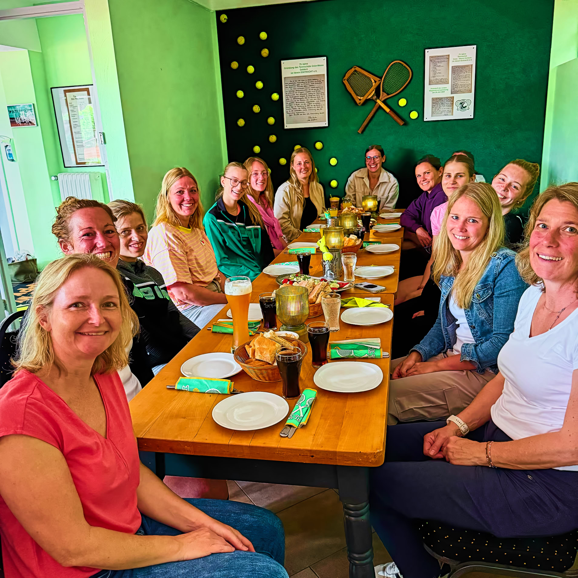Eine Gruppe von Menschen sitzt um einen langen Tisch in einem Raum mit gr&uuml;nen W&auml;nden, auf denen Tennisschl&auml;ger h&auml;ngen. Auf dem Tisch stehen Teller und Getr&auml;nke.