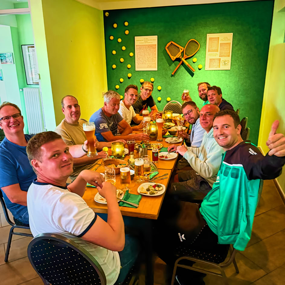 Eine Gruppe von Personen sitzt um einen Tisch in einem Restaurant, l&auml;chelnd und mit Getr&auml;nken und Essen vor sich. Die W&auml;nde sind gr&uuml;n dekoriert, mit Tennisschl&auml;gern und Tennisb&auml;llen an der Wand.