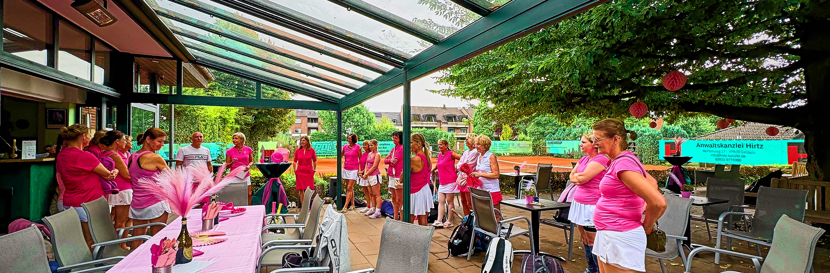 Eine Gruppe von Menschen in pinker Kleidung steht unter einem Glasdach in einem Au&szlig;enbereich mit Tischen und St&uuml;hlen.