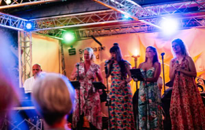 Eine Gruppe von f&uuml;nf Personen auf einer B&uuml;hne, vier singen in Mikrofone, eine spielt Keyboard, bunte Beleuchtung im Hintergrund.