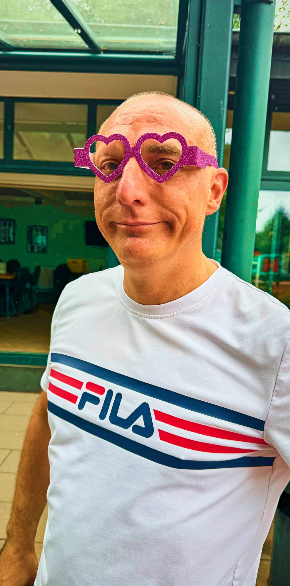 Eine Person mit Glatze tr&auml;gt eine wei&szlig;e T-Shirt mit dem Aufdruck "FILA" und eine herzf&ouml;rmige, rosa Brille. Im Hintergrund sind ein Geb&auml;ude mit gro&szlig;en Fenstern und Pflanzen zu sehen.