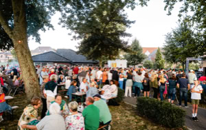 Eine gro&szlig;e Menschenmenge versammelt sich im Freien bei einem Fest oder Event, umgeben von B&auml;umen und Geb&auml;uden im Hintergrund.