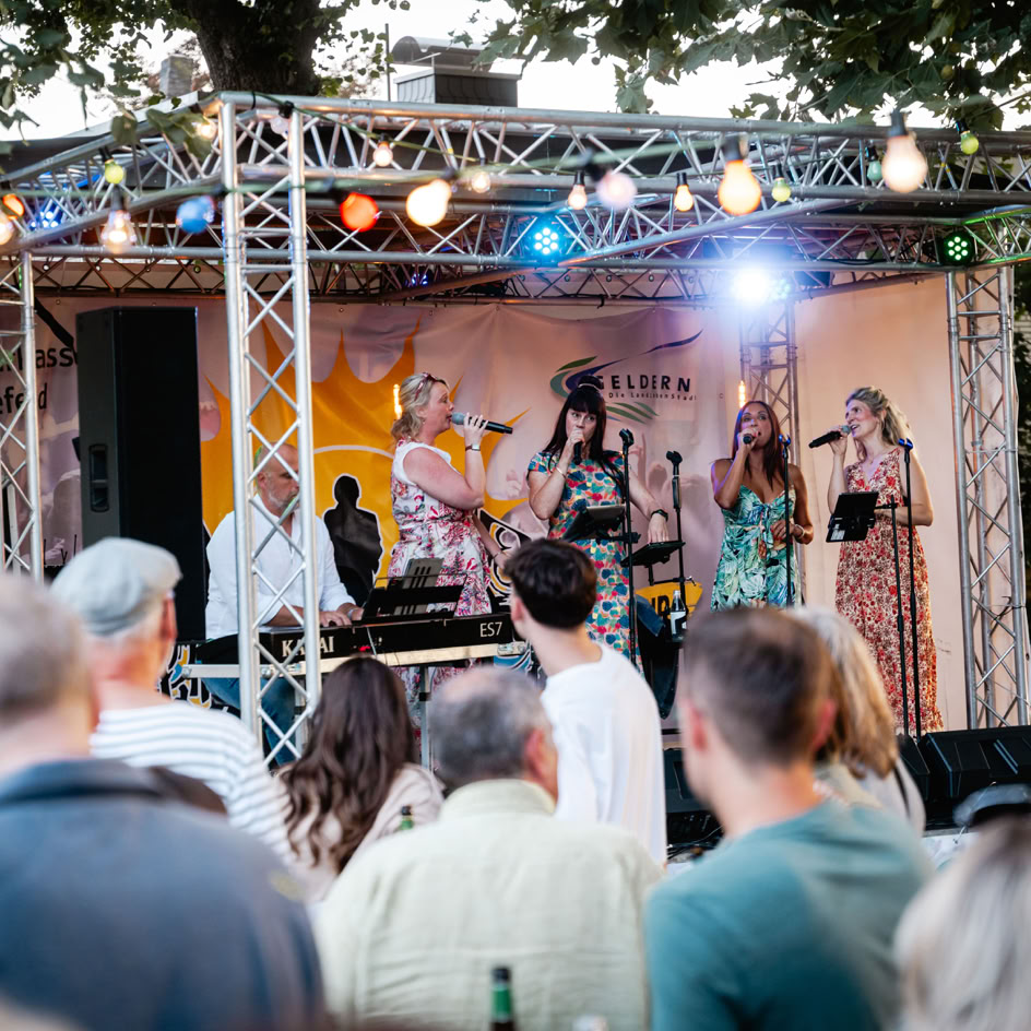 Eine Band spielt auf einer B&uuml;hne im Freien, umgeben von B&auml;umen und bunten Lichtern, w&auml;hrend Menschen im Vordergrund zuschauen.