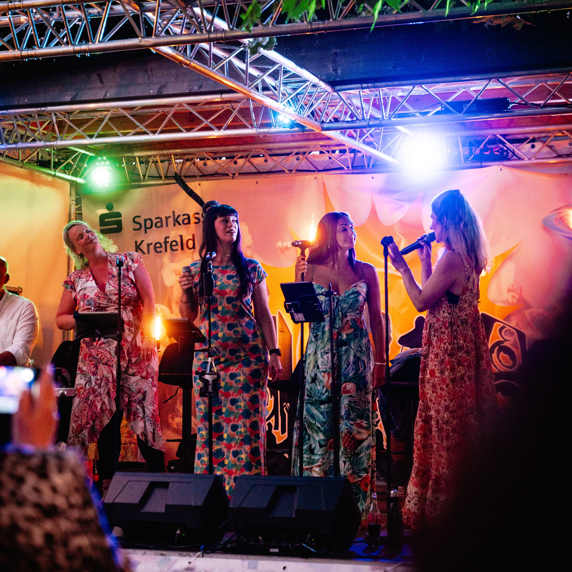 Vier Personen stehen auf einer B&uuml;hne und singen in Mikrofone, umgeben von bunten Lichtern und Dekorationen.
