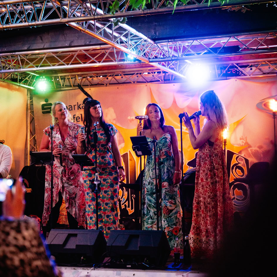 Vier Personen singen auf einer B&uuml;hne unter bunten Lichtern, umgeben von B&auml;umen und einem Banner im Hintergrund.