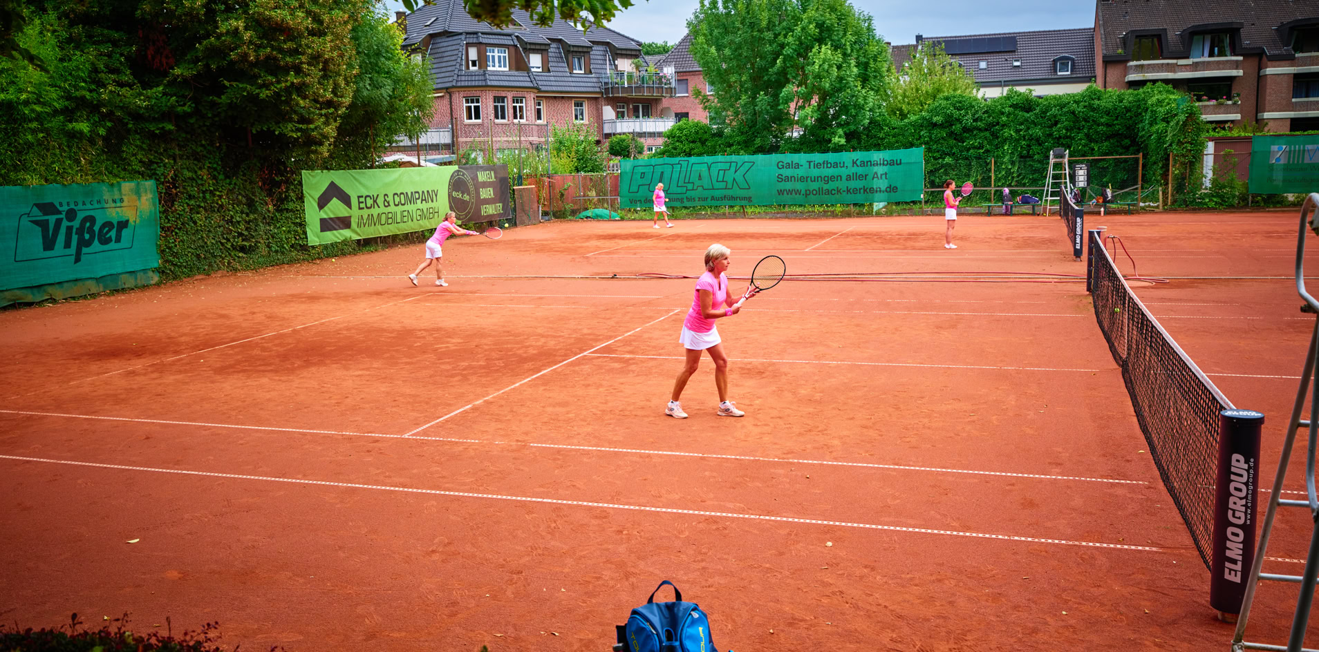 Tennisplatz mit mehreren Personen, die Tennis spielen; im Vordergrund sind Tennistaschen zu sehen, im Hintergrund B&auml;ume und Geb&auml;ude.
