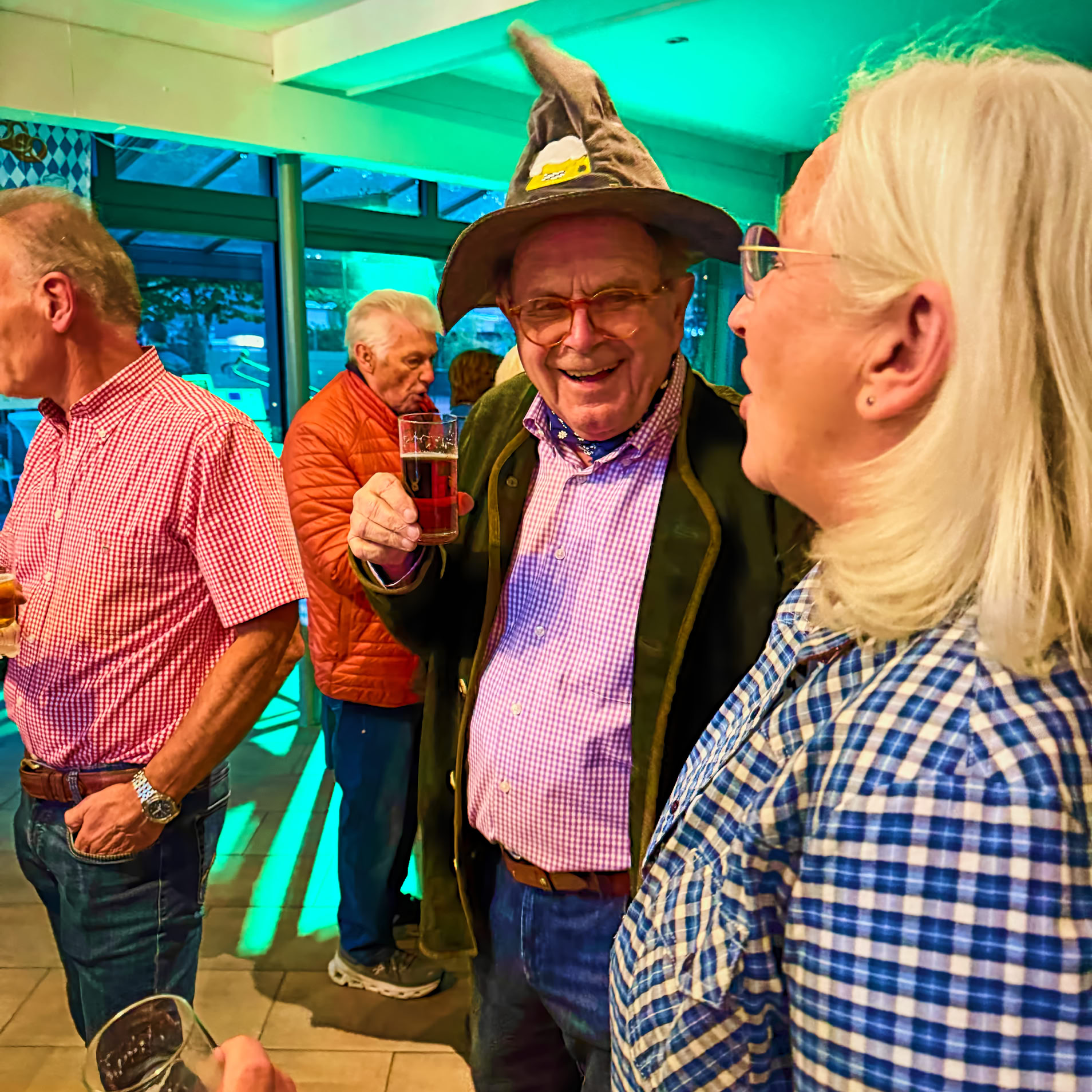 Eine Gruppe von &auml;lteren Menschen in einem Raum, einige halten Getr&auml;nke. Eine Person tr&auml;gt einen Hut mit Federn.