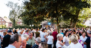 Eine gro&szlig;e Menschenmenge versammelt sich im Freien bei Tageslicht, umgeben von B&auml;umen und Geb&auml;uden im Hintergrund.