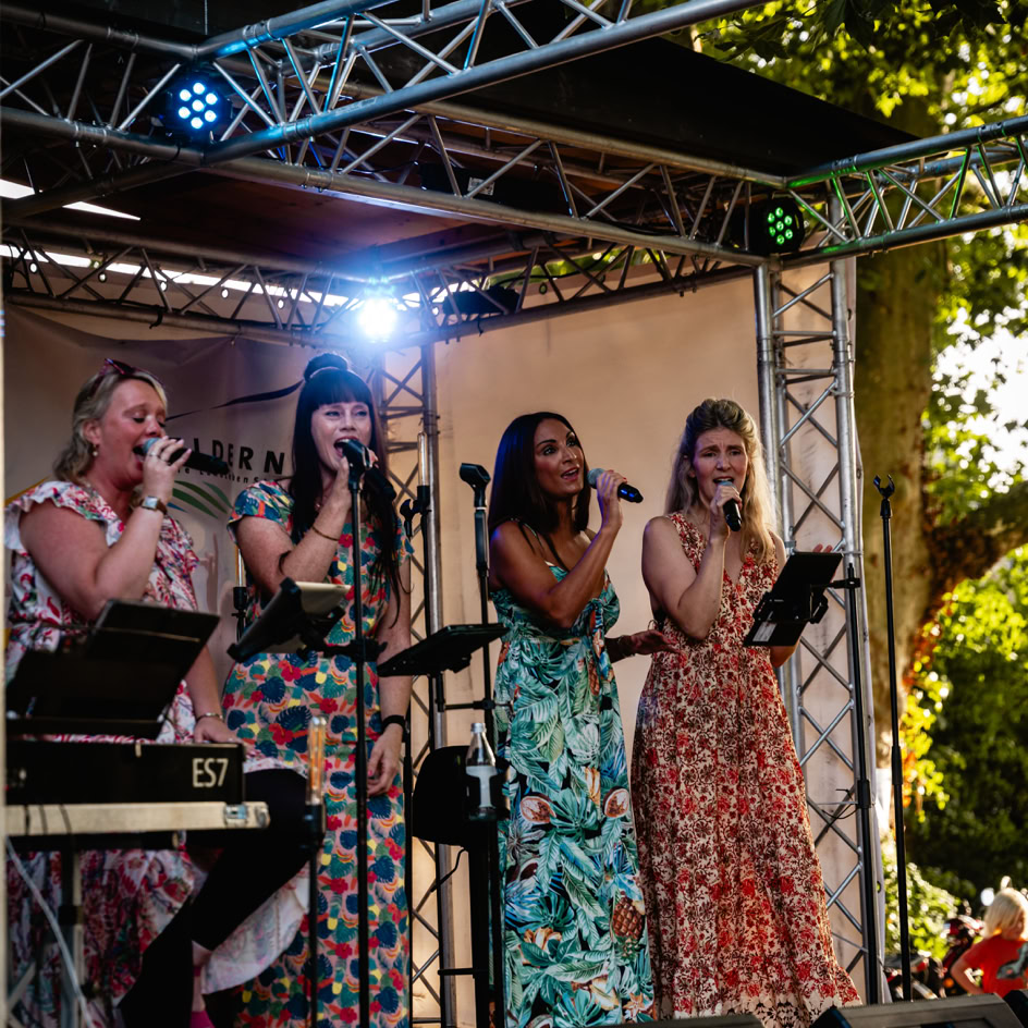 Vier Frauen singen auf einer B&uuml;hne im Freien, umgeben von B&auml;umen und Lichtern. Sie tragen bunte Kleider und halten Mikrofone.