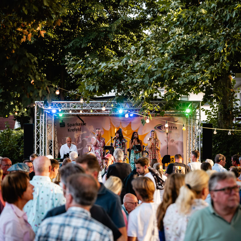 Eine Menschenmenge steht vor einer B&uuml;hne im Freien, auf der eine Band unter bunten Lichtern auftritt; umgeben von B&auml;umen.