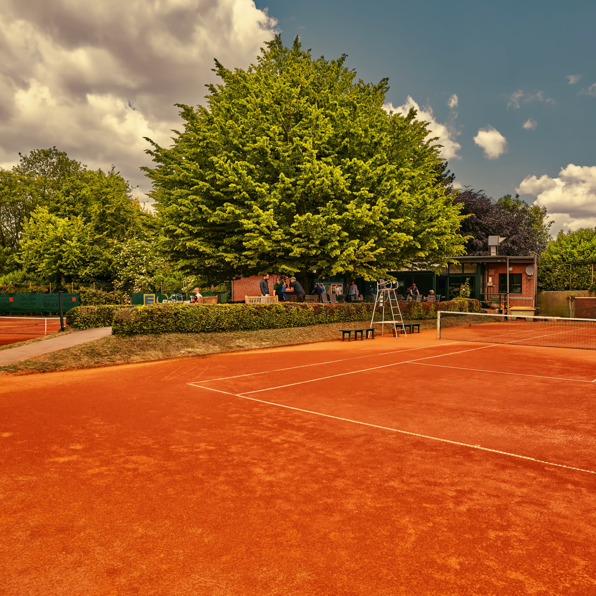 Ein roter Sandtennisplatz mit einem Baum und einem Geb&auml;ude im Hintergrund, umgeben von gr&uuml;nen B&auml;umen und einem blauen Himmel mit Wolken.