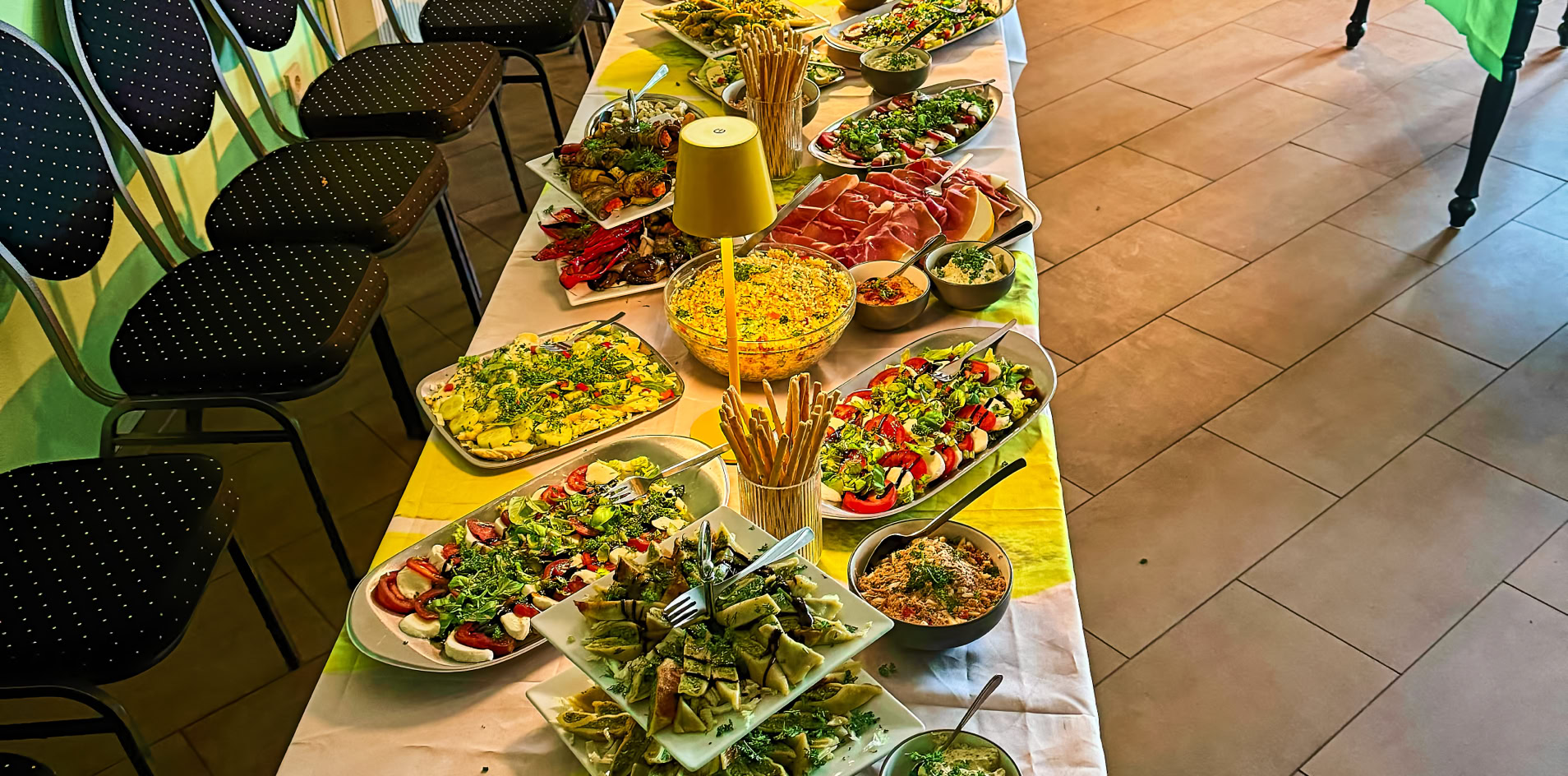 Langer Tisch mit wei&szlig;er Tischdecke, darauf verschiedene Speisen und Dekorationen; im Hintergrund eine gr&uuml;ne Wand und eine Bar.