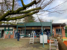 Geb&auml;ude mit Glasfront, mehreren Leitern und Personen, die im Au&szlig;enbereich arbeiten; im Vordergrund ein gro&szlig;er Baum.