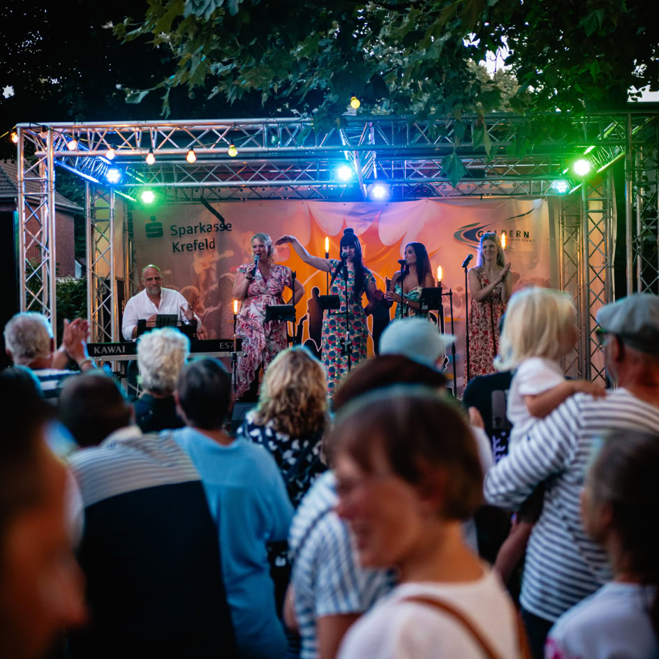 Eine Menschenmenge steht vor einer B&uuml;hne im Freien, auf der eine Band unter bunten Lichtern spielt.