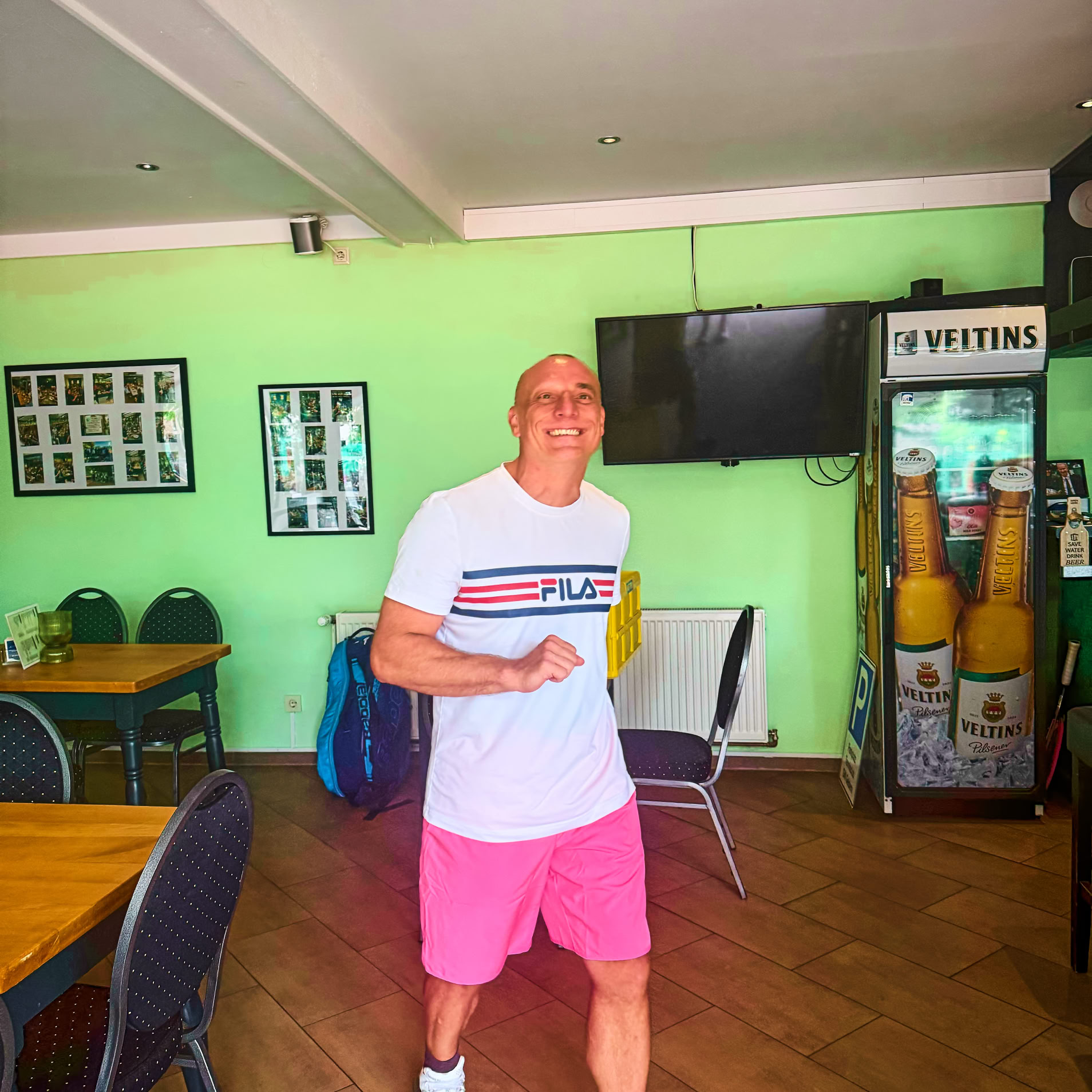 Ein Mann in einem wei&szlig;en T-Shirt und pinken Shorts steht in einem Raum mit gr&uuml;nen W&auml;nden, Tischen und St&uuml;hlen. Im Hintergrund sind ein Fernseher und ein K&uuml;hlschrank zu sehen.