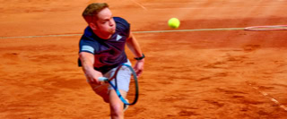 Person spielt Tennis auf einem Sandplatz, h&auml;lt einen Schl&auml;ger und schaut nach oben, w&auml;hrend ein Ball in der Luft schwebt.