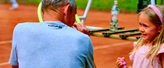 Ein Mann in einem grauen T-Shirt spricht mit einem M&auml;dchen mit rosa Haarband auf einem Tennisplatz. Im Hintergrund sind unscharfe Personen und ein Tennisnetz zu sehen.