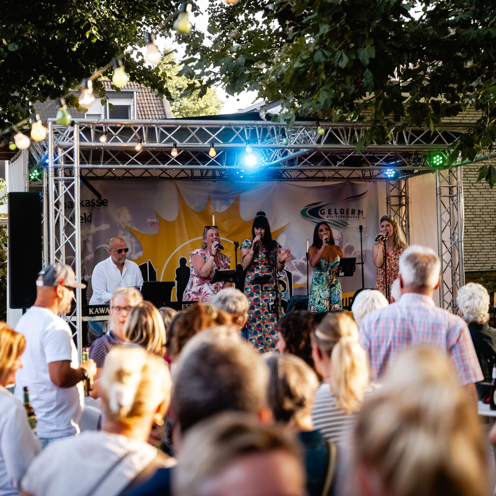 Eine Gruppe von Menschen steht vor einer B&uuml;hne im Freien, auf der mehrere Musiker unter einem Ger&uuml;st mit Lichtern auftreten. B&auml;ume und Lichterketten sind im Hintergrund zu sehen.