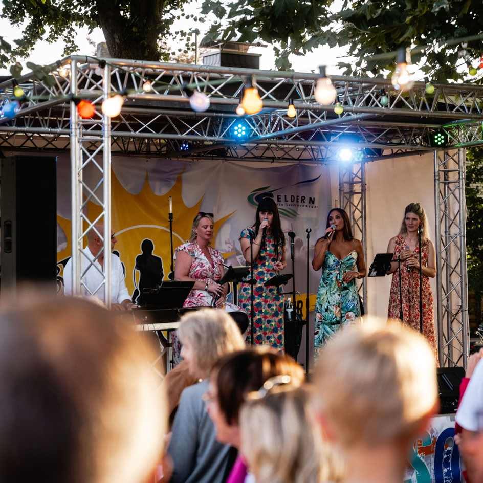 Eine Gruppe von Menschen steht auf einer B&uuml;hne im Freien und singt oder spielt Musik, w&auml;hrend ein Publikum zuschaut. Die B&uuml;hne ist mit Lichtern dekoriert.