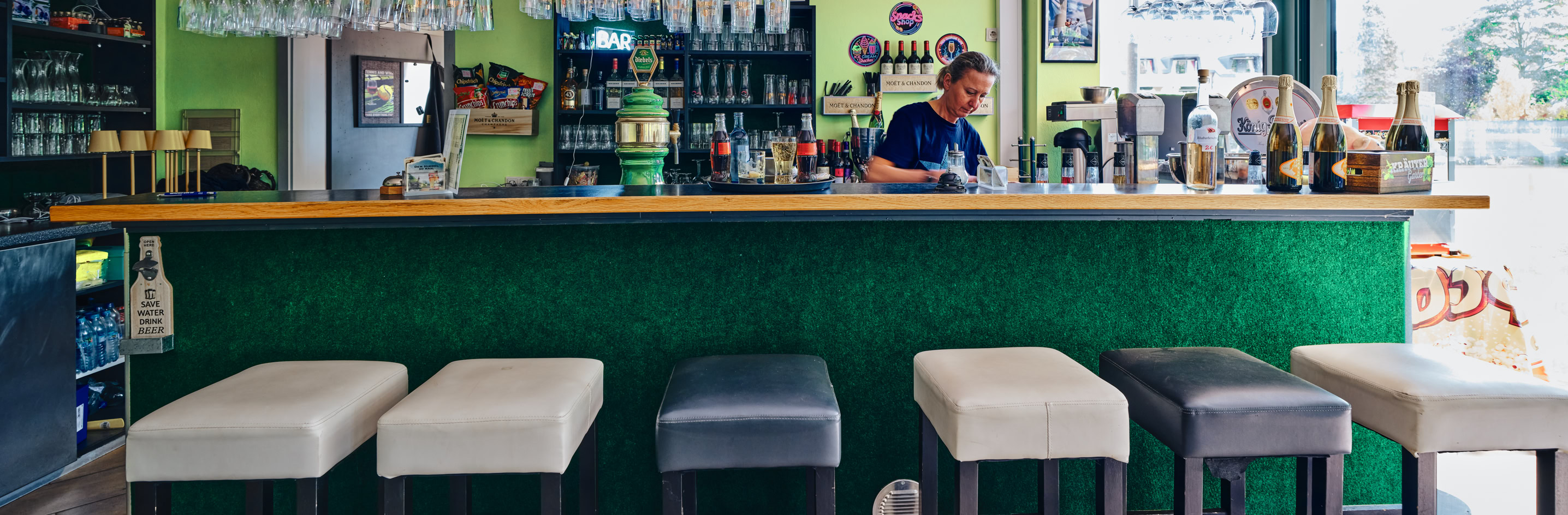 Bar mit gr&uuml;ner Theke, f&uuml;nf Barhocker davor, Gl&auml;ser und Flaschen im Hintergrund, Person hinter der Theke.