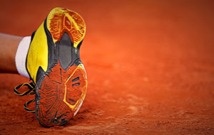 Ein gelber Sportschuh mit schwarzer Sohle liegt auf einem roten Sandplatz.