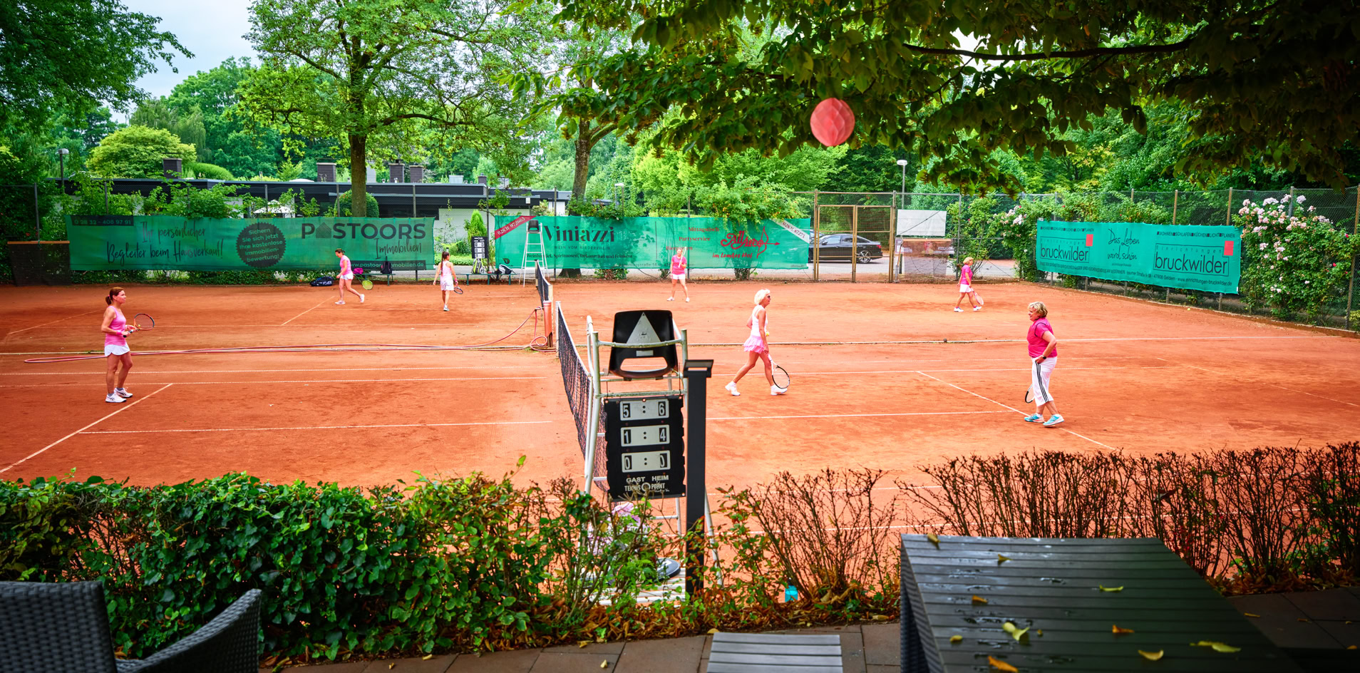 Tennisplatz mit mehreren Personen beim Spielen, umgeben von B&auml;umen und gr&uuml;nen Z&auml;unen, im Vordergrund eine Terrasse mit St&uuml;hlen und Tischen.