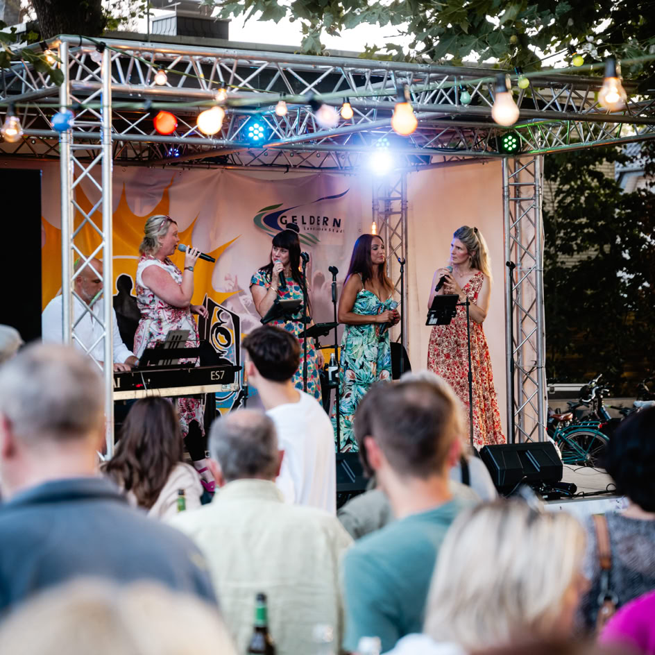 Eine Gruppe von Menschen steht vor einer B&uuml;hne im Freien, auf der mehrere Personen in bunten Kost&uuml;men auftreten; bunte Lichter und B&auml;ume im Hintergrund.