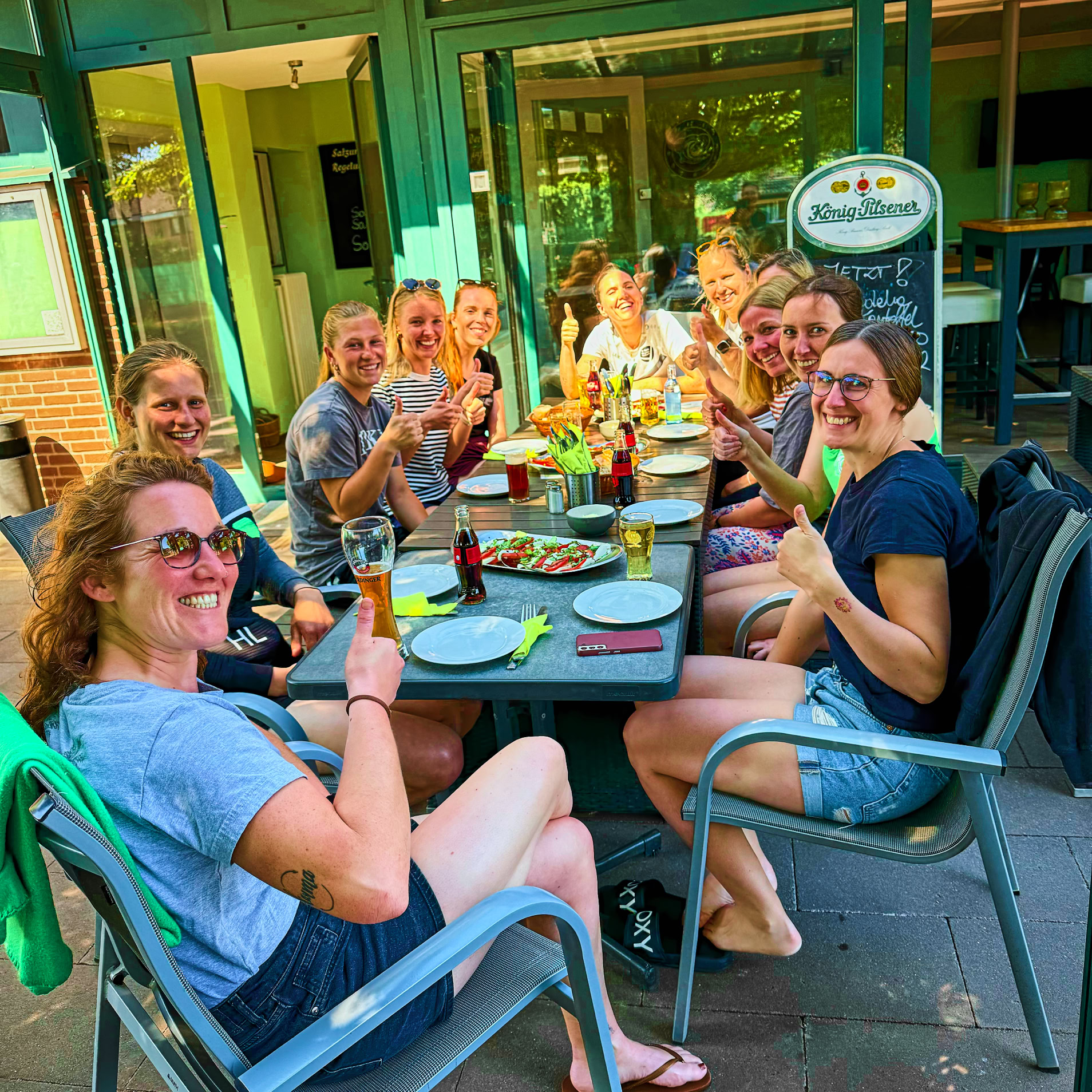 Eine Gruppe von Menschen sitzt an einem Tisch im Freien, l&auml;chelt und hebt Gl&auml;ser.