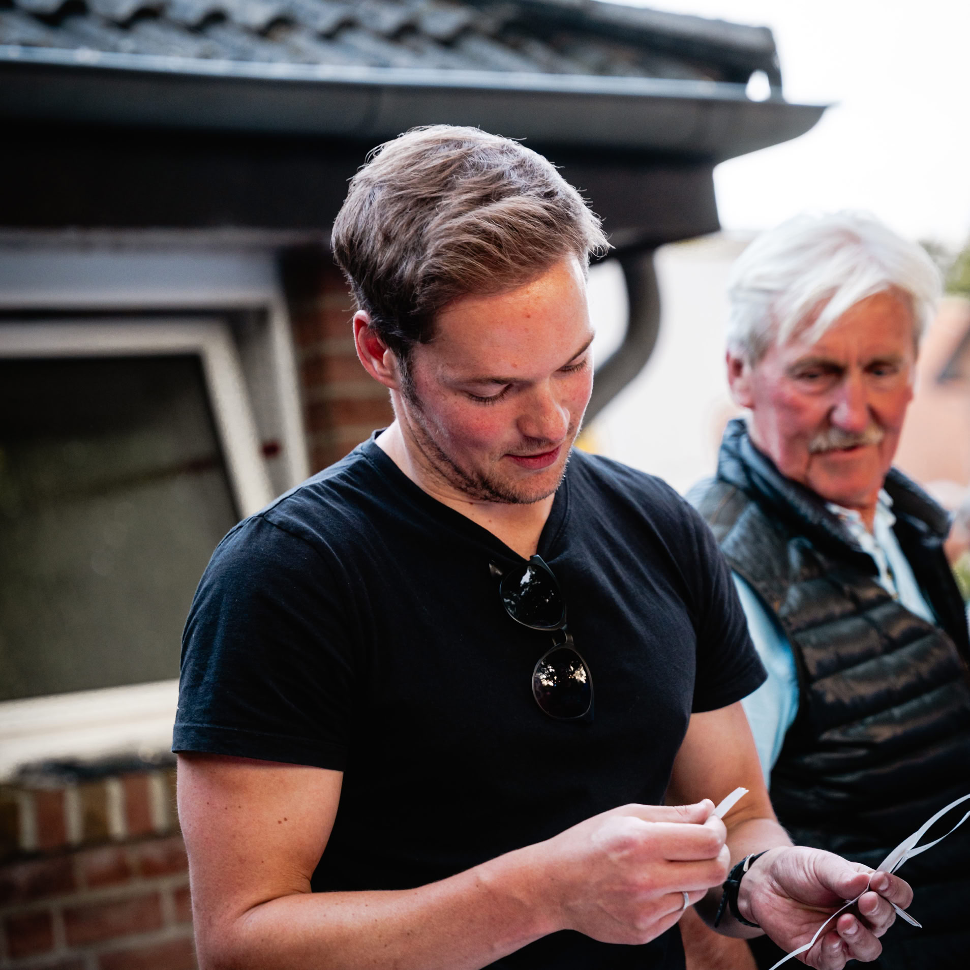 Ein Mann in einem schwarzen T-Shirt h&auml;lt etwas in den H&auml;nden, w&auml;hrend ein &auml;lterer Mann daneben steht. Im Hintergrund ist ein Hausdach und ein Fenster zu sehen.
