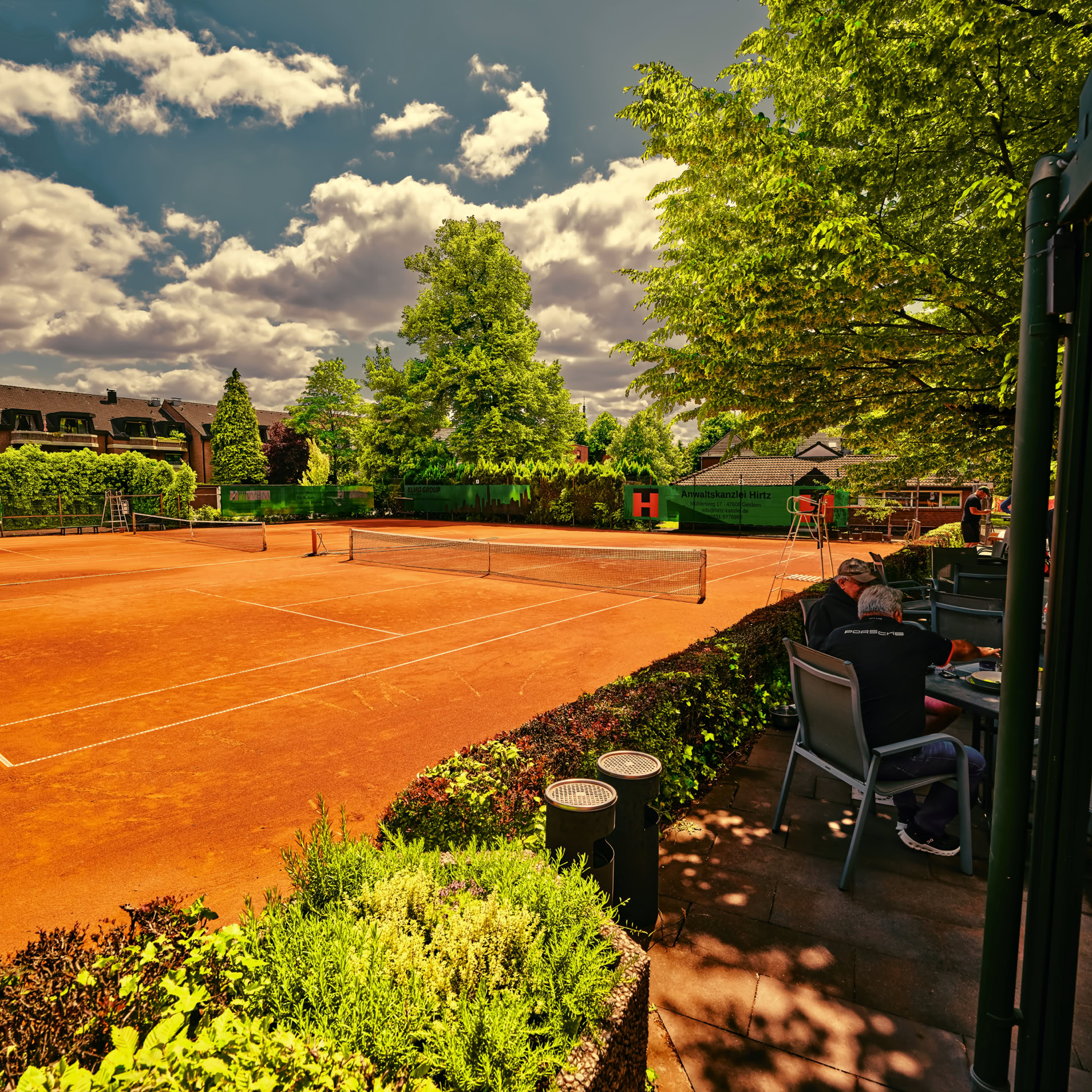 Tennisplatz mit rotem Sand, umgeben von B&auml;umen und B&uuml;schen, im Hintergrund Geb&auml;ude; im Vordergrund sitzen Menschen an einem Tisch.