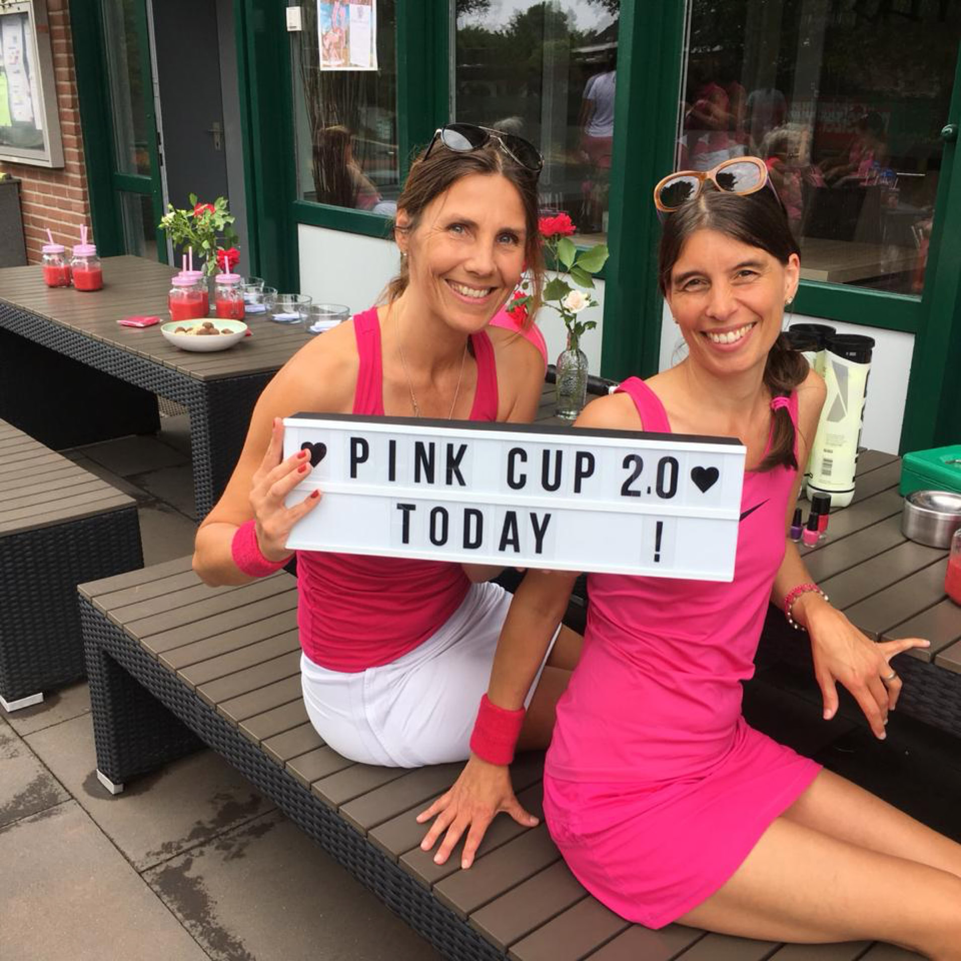 Zwei Frauen in pinker Sportkleidung sitzen auf einer Bank und halten ein Schild mit der Aufschrift "Pink Cup 20 Today". Im Hintergrund sind Tische mit Getr&auml;nken und ein Geb&auml;ude mit Fenstern zu sehen.