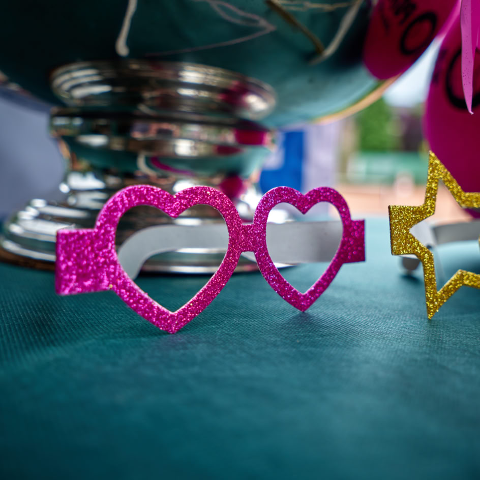Herzf&ouml;rmige, pinke Brille auf einem Tisch vor einem silbernen Pokal, daneben eine sternf&ouml;rmige, goldene Brille.
